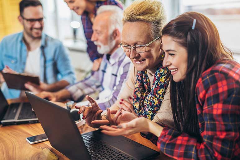 Photograph of diverse group of elderly and young people sitting at a table working with laptop computers.